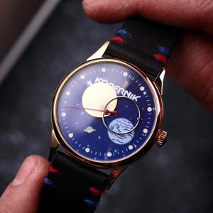 May include: A gold-toned wristwatch with a black leather band and a blue face featuring a celestial design. The watch face displays a moon, planets, and stars. The band has red and blue stitching.