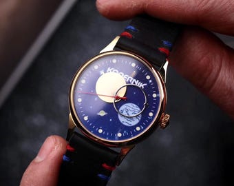 May include: A gold-toned wristwatch with a black leather band and a blue face featuring a celestial design. The watch face displays a moon, planets, and stars. The band has red and blue stitching.