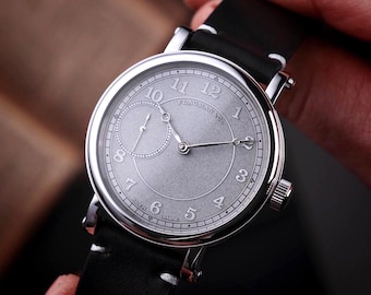 May include: A silver-toned wristwatch with a black leather band. The watch face has a textured, light gray finish with black numerals and hands. A smaller subdial is positioned at the 9 o'clock position. The watch is held by a person's hand.