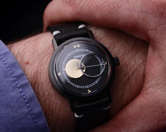 May include: A black wristwatch with a unique dial design featuring a gold-colored circle and a black crescent shape. The watch has a black leather band and is worn on a wrist with a blue shirt sleeve visible.