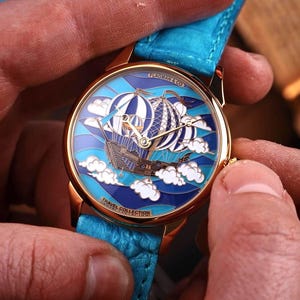 Op de afbeelding: Goudkleurig polshorloge met een blauwe leren band. De wijzerplaat toont een gedetailleerde illustratie van luchtballonnen en een schip dat door de wolken vaart. Het horloge maakt deel uit van de "Travel Collection".