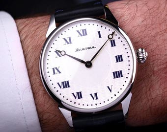 Könnte beinhalten: Nahaufnahme einer Armbanduhr mit weißem Zifferblatt und schwarzen Zeigern. Die Uhr hat schwarze römische Ziffern und ein schwarzes Lederarmband. Auf dem Zifferblatt steht das Wort "Флагман". Die Uhr befindet sich an einem Handgelenk.
