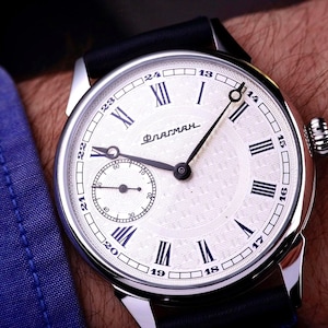 Könnte beinhalten: Silberne Armbanduhr mit weißem Zifferblatt, schwarzen römischen Ziffern und einer kleinen Sekundenanzeige. Die Uhr hat schwarze Zeiger und ein schwarzes Lederarmband. Das Wort "Флагман" ist auf dem Zifferblatt aufgedruckt.