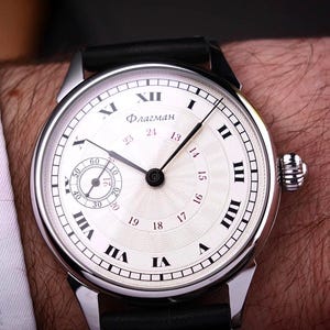 May include: A silver wristwatch with a white face and black Roman numerals. The watch has a small seconds dial and a black leather band. The word "Флагман" is printed on the watch face. The watch is on a person's wrist.