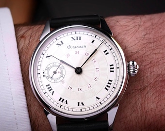 Könnte beinhalten: Eine silberne Armbanduhr mit weißem Zifferblatt und schwarzen römischen Ziffern. Die Uhr hat ein kleines Sekunden-Zifferblatt und ein schwarzes Lederarmband. Das Wort "Флагман" ist auf dem Zifferblatt aufgedruckt. Die Uhr befindet sich am Handgelenk einer Person.