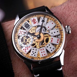 May include: A silver wristwatch with a black leather band. The watch face features a gold-colored inner mechanism with playing card designs and visible gears. The watch is on a person's wrist.
