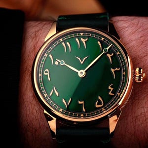 Può includere: Un orologio color oro con quadrante verde e numeri arabi. L'orologio ha lancette dorate e un cinturino in pelle verde scuro. L'orologio è su un polso.