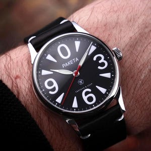 Könnte beinhalten: Eine schwarze Armbanduhr mit silbernem Gehäuse und schwarzem Lederarmband. Das Zifferblatt hat große weiße Ziffern, weiße Zeiger und das Wort "PAKETA". Der Sekundenzeiger ist rot.