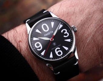 May include: A black wristwatch with a silver case and a black leather band. The watch face features large white numerals, white hands, and the word "PAKETA". The second hand is red.