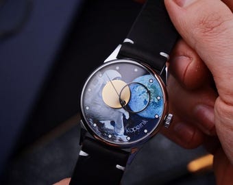 May include: A silver wristwatch with a black leather band. The watch face features a celestial design with a light blue and white planet, a gold circle, and the word "Kopernik". The watch is held by a person.