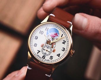 May include: A gold-toned wristwatch with a white face featuring a cartoon illustration and black numerals. The watch has a brown leather strap. The watch is being held by a person.