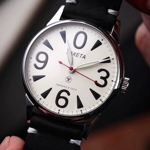 May include: A silver wristwatch with a white face, black numerals, and black hands. The watch has a black leather strap with white stitching. The watch face has the word "Raketa" and the text "Made in USSR".