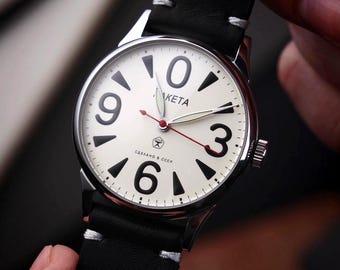 May include: A silver wristwatch with a white face, black numerals, and black hands. The watch has a black leather band with white stitching. The watch face has the word "Raketa" and the text "Made in USSR".