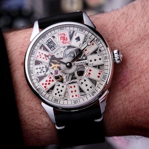 May include: A silver wristwatch with a black leather band. The watch face features a playing card design with visible internal gears. The watch hands point to the time. The word "BARMAN" is visible on the watch face.