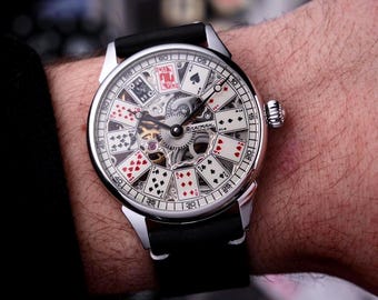 May include: A silver wristwatch with a black leather band. The watch face features a playing card design with visible internal gears. The watch hands point to the time. The word "BARMAN" is visible on the watch face.