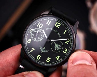 Könnte beinhalten: Schwarze Armbanduhr mit schwarzem Lederarmband. Das Zifferblatt ist schwarz mit weißen Ziffern und Zeigern und dem Text "Штурманские". Verfügt über eine kleine Sekundenanzeige und eine Flugzeuggrafik. Die Uhr wird von einer Person gehalten.