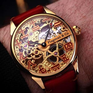 Op de afbeelding: Goudkleurig polshorloge met een rode leren band. De wijzerplaat heeft ingewikkelde open-werk details met rode accenten en de tekst "Flagman & Co." Het horloge zit om een pols.