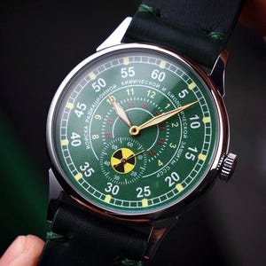 May include: A green-faced wristwatch with gold hands and a black leather strap. The watch face features a radiation symbol and text in a foreign language. The watch has a silver casing and a crown on the side.