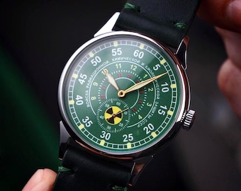 May include: A green-faced wristwatch with gold hands and a black leather strap. The watch face features a radiation symbol and text in a foreign language. The watch has a silver casing and a crown on the side.