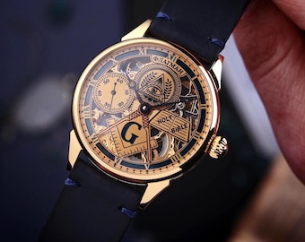 May include: A gold-toned wristwatch with a black leather band. The watch face features intricate Masonic symbols, including an all-seeing eye, a compass, and the letter "G". The watch has two sub-dials and Roman numeral hour markers.