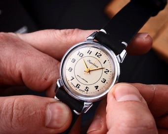 Könnte beinhalten: Eine Vintage-Armbanduhr mit cremefarbenem Zifferblatt, schwarzen Ziffern und goldenen Zeigern. Die Uhr hat ein silberfarbenes Gehäuse und ein schwarzes Lederarmband. Auf dem Zifferblatt steht "Zaria". Die Uhr wird von zwei Händen gehalten.