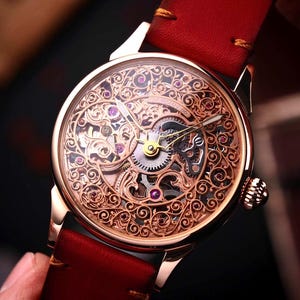 Op de afbeelding: Een goudkleurig skeleton horloge met een ingewikkeld filigraan ontwerp op de wijzerplaat. Het horloge heeft zichtbare tandwielen en juwelen, gezet tegen een rode leren band. Het horloge wordt door een hand vastgehouden.