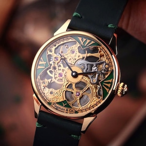 Może przedstawiać: Zegarek szkieletowy w kolorze złotym z zielono-złotą tarczą i czarnym skórzanym paskiem. Tarcza zegarka posiada rzymskie cyfry i skomplikowane detale mechaniczne. Zegarek jest marki "Flagman & Co."