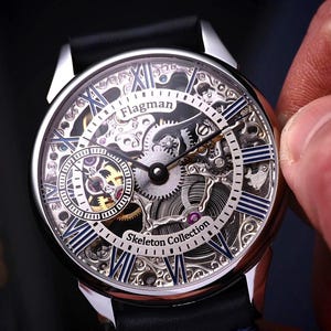 Op de afbeelding: Een zilveren skeleton horloge met een zwarte leren band. De wijzerplaat toont zichtbare tandwielen, Romeinse cijfers en de tekst "Flagman Skeleton Collection". Het horloge wordt door een persoon vastgehouden.