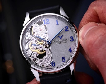Könnte beinhalten: Silberfarbene Armbanduhr mit schwarzem Lederarmband. Das Zifferblatt ist geteilt, wobei eine Seite das komplizierte Uhrwerk und die andere ein hellgraues Zifferblatt mit blauen Ziffern und Zeigern zeigt. Die Uhr wird eingestellt.