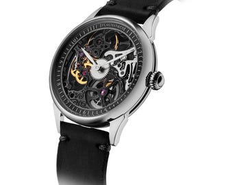 Könnte beinhalten: Eine silberfarbene Armbanduhr mit schwarzem Lederarmband. Das skelettierte Zifferblatt zeigt das Innenleben mit goldenen und rubinroten Akzenten. Die Uhr trägt den Schriftzug "FLAGMAN & CO."