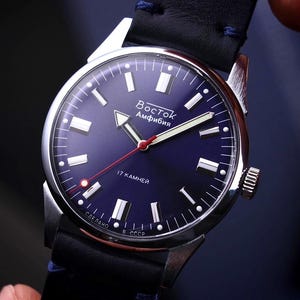 May include: A close-up of a blue-faced Vostok Amphibia watch with a silver case and black leather strap. The watch face features white markers, a red second hand, and the text "Восток Amphibia".
