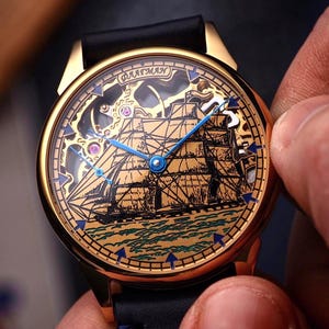 Op de afbeelding: Een goudkleurig horloge met een zwarte leren band. De wijzerplaat van het horloge toont een gedetailleerde illustratie van een schip op zee, met blauwe wijzers en accenten. Het horloge wordt door een persoon vastgehouden.