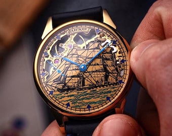 May include: A gold-toned wristwatch with a black leather band. The watch face features a detailed illustration of a ship on the sea, with blue hands and accents. The watch is being held by a person.