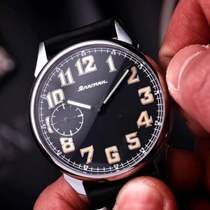 May include: A black wristwatch with a silver bezel and a black leather strap. The watch face has large, cream-coloured numerals and a smaller sub-dial. The watch is being adjusted by a person's hand.