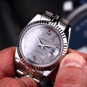 Op de afbeelding: Een zilveren polshorloge met een grijze wijzerplaat met illustraties van verschillende poses. De wijzerplaat bevat de tekst "FLAGMAN & CO" en "KAMASUTRA". Het horloge heeft een zilveren band en een getextureerde bezel.