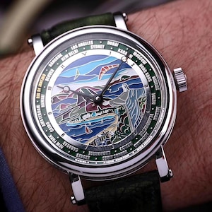 Op de afbeelding: Een zilveren polshorloge met een groene leren band. De wijzerplaat van het horloge heeft een kleurrijk landschapsontwerp met een berg-, oceaan- en stadsgezicht. De bezel toont stadsnamen. Het horloge zit om een pols.