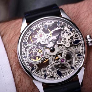 May include: A silver Flagman skeleton watch with a black leather strap. The watch face displays intricate gears and mechanisms, with silver, gold, and purple details. The watch is on a wrist, with a white shirt sleeve in the background.