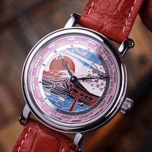 Op de afbeelding: Een zilveren polshorloge met een rode leren band. De wijzerplaat toont een Japans landschap met een rode zon, een berg en een traditionele poort. Het horloge heeft een roze en witte rand met stadsnamen.