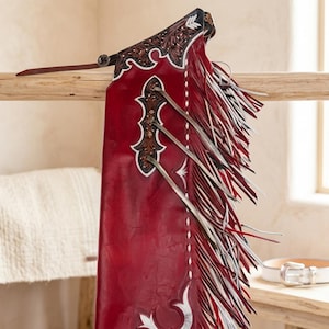 May include: A deep red leather chap featuring intricate dark brown floral tooling at the top and side panel. The garment includes long, layered fringe in red, white, and grey, accented with white stitching and a thin adjustable leather strap at the top.