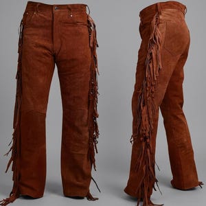 May include: A pair of brown suede leather trousers featuring long, dangling fringe detailing along the outer seams of both legs. The trousers have a standard waistband with belt loops, a button closure, and a straight-leg cut shown from two angles.