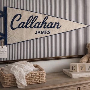 Op de afbeelding: Een ingelijste wimpel met de naam "Callahan JAMES" in marineblauw schrift. De crèmekleurige wimpel hangt tegen een gestreepte achtergrond. Een teddybeer, houten blokken en een rieten mand zijn ook aanwezig.