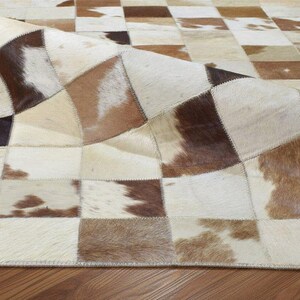 May include: A patchwork rug with a cowhide design. The rug features squares of varying shades of brown, cream, and tan. The rug is partially rolled up, revealing the underside and the stitched edges.