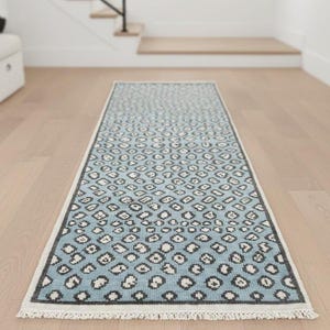 May include: A rectangular, light blue rug with a repeating pattern of black and white shapes. The rug has a white fringe on the short ends and a dark gray border. The rug is placed on a light wood floor.