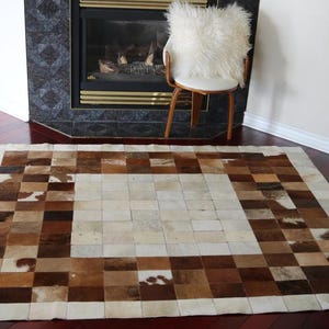 Könnte beinhalten: Rechteckiger Kuhfellteppich mit Patchwork-Design. Der Teppich hat Quadrate in verschiedenen Braun-, Beige- und Cremetönen. Der Teppich liegt auf einem dunklen Holzboden, mit einem Kamin und einem Stuhl im Hintergrund.