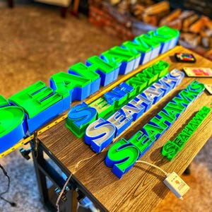 Puede incluir: Varios letreros "Seahawks" impresos en 3D en varios tamaños y estilos, con colores verde, azul y plateado. Los letreros se exhiben sobre una superficie de madera, con una cinta métrica y pequeños componentes electrónicos.