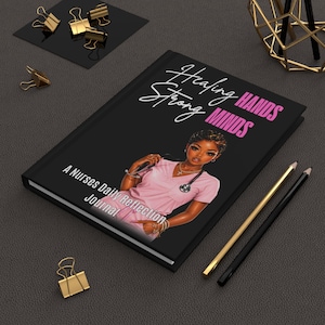 May include: Black journal with the text "Healing Hands Strong Minds" and "A Nurses Daily Reflection Journal". The cover features an illustration of a nurse in pink scrubs. Gold and black pencils and gold binder clips are also visible.