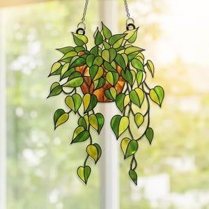 May include: Stained glass hanging planter with green and yellow leaves. The plant is in a copper-colored pot and hangs from a silver chain. The leaves have a heart shape. The background is a blurred view of a window and trees.