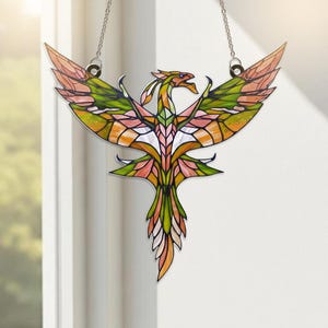 May include: A stained glass phoenix with outstretched wings, suspended by a chain. The bird features a vibrant color palette of orange, green, pink, and white. The artwork is displayed against a white background, near a window.