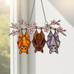 K&ouml;nnte beinhalten: Ein Fensterbehang aus Buntglas mit drei Flederm&auml;usen, die an einem Ast mit rosa Bl&uuml;ten h&auml;ngen. Die Flederm&auml;use sind orange, braun und lila. Der Ast ist hellbraun und die Bl&uuml;ten sind rosa. Aufgeh&auml;ngt an einer silbernen Kette.