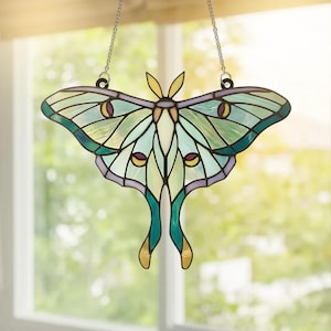 May include: A stained glass suncatcher shaped like a luna moth, featuring translucent panels in shades of mint green, pale yellow, and lavender, outlined in dark metal solder. The moth has two long, trailing lower wings and a silver chain.